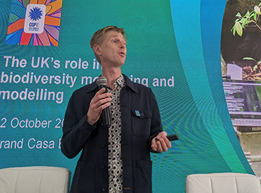 Dr Nick Isaac speaking in front of a large screen at a COP16 event