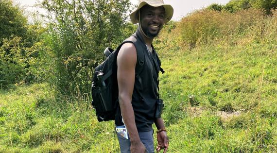 Joseph Ibrahim during his visit Joseph Ibrahim stands in a grassy area wearing jeans, a black vest, a beige hat and a rucksack