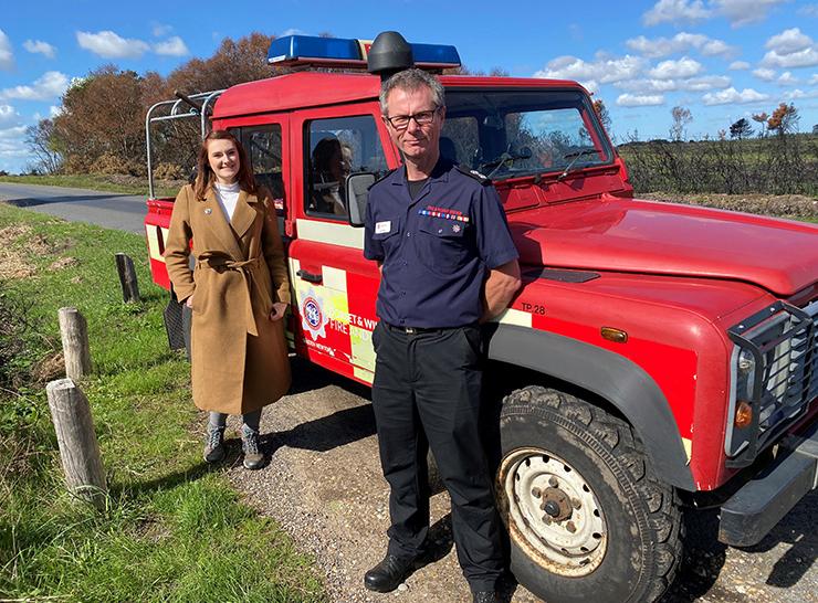 Two people stand in front of a red land rover with a fire brigade crest and blue lights