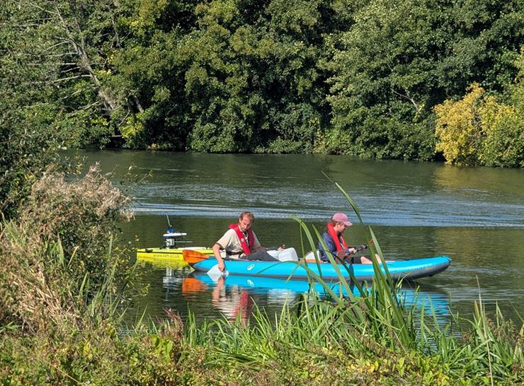 Two people sit in a kayak on a river beside a remote controlled survey boat